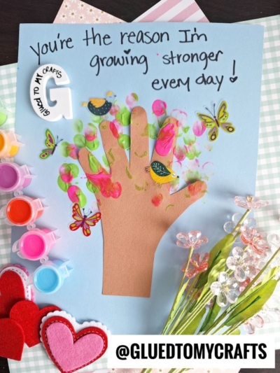 A handmade card features a brown paper cutout of a hand decorated with paint, butterfly, and bird stickers on blue paper, surrounded by paint pots, faux flowers, hearts, and the text You’re the reason I’m growing stronger every day!.