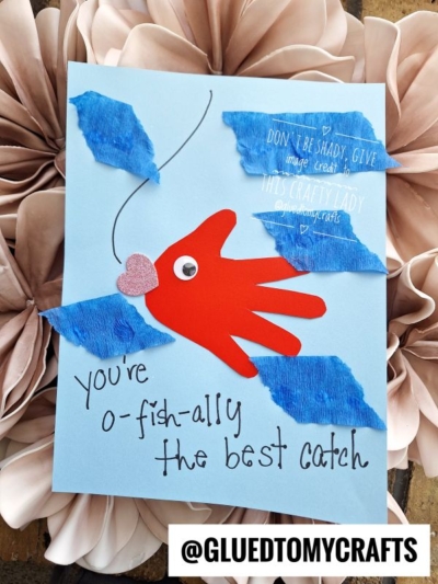 A handmade card shows a red fish made from a handprint cutout, blue tissue paper water, and a heart-shaped pink mouth. It reads Youre o-fish-ally the best catch on a light blue background.