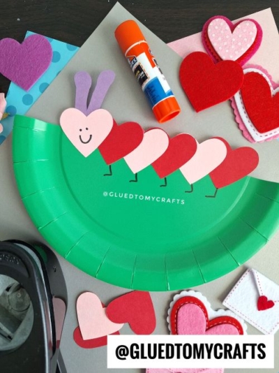 A Valentine's Day caterpillar craft made from a green paper plate cut in half, with alternating red and pink heart-shaped segments forming the body. The head is a pink heart with a smiley face and purple antennae, and black legs are drawn on the plate. Surrounding the craft are felt hearts, a glue stick, and a hole punch, all arranged on a gray surface with patterned papers.