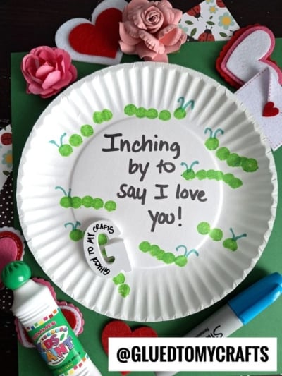 A paper plate decorated with green caterpillars made from paint dots surrounds the handwritten message, “Inching by to say I love you!” Craft supplies and heart decorations are scattered around.