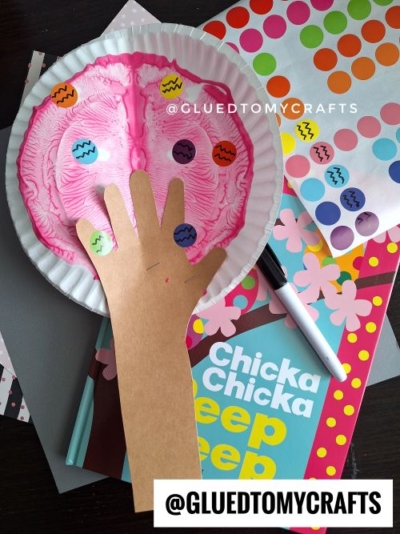 A children's craft featuring a cherry blossom tree made from a pink paint-splattered paper plate as the foliage and a brown handprint cutout as the trunk. The tree is decorated with colorful dot stickers resembling Easter eggs. The craft is surrounded by a children's book titled "Chicka Chicka Peep Peep," a black marker, and patterned papers, with the watermark "@GLUEDTOMYCRAFTS" visible.