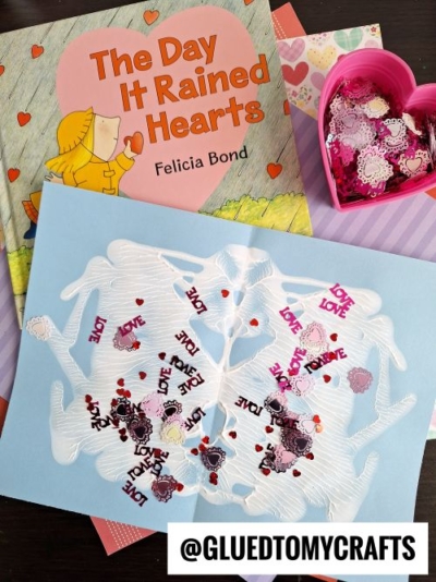 A flat lay of Valentine's Day-themed craft supplies and a children's book titled "The Day It Rained Hearts" by Felicia Bond. The craft features a light blue paper with a white glue or paint design, adorned with red heart confetti and "LOVE" letter sequins. A pink heart-shaped container filled with similar embellishments is nearby. The background includes colorful patterned papers in pink, red, and purple tones.