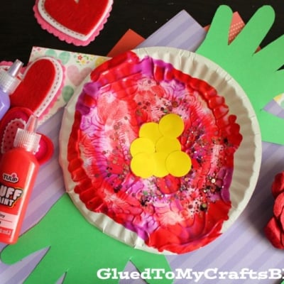 A colorful paper craft featuring a red and pink painted paper plate with yellow circles in the center, green handprint cutouts as leaves, surrounded by craft supplies, heart decorations, and paint bottles.