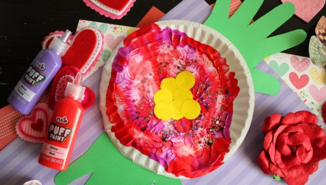 A colorful paper craft featuring a red and pink painted paper plate with yellow circles in the center, green handprint cutouts as leaves, surrounded by craft supplies, heart decorations, and paint bottles.
