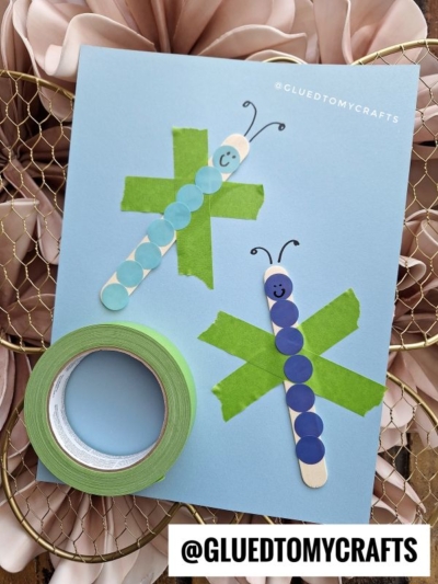A handmade card with two caterpillars made from colored paper dots on popsicle sticks, each with a smiley face, on a blue background with green tape Xs. A roll of green tape is nearby.