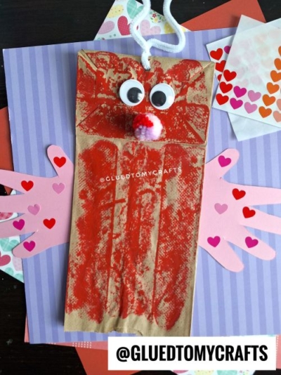 A Valentine's Day-themed craft made from a brown paper bag, decorated with red paint, googly eyes, a pom-pom nose, and pink paper hands adorned with heart stickers. The craft is set against a colorful background of patterned papers featuring hearts and stripes.