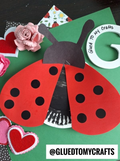 A handmade ladybug craft with red wings and black spots, surrounded by paper flowers and heart decorations on a green background. The username @GLUEDTOMYCRAFTS is at the bottom.