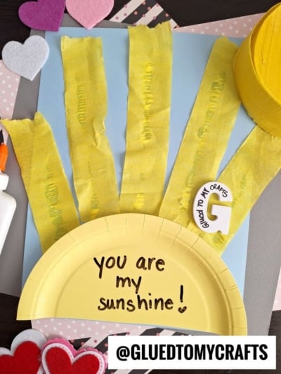 A craft project made with a yellow paper plate, yellow tissue strips, glue, gray paper, and felt hearts. The plate says You are my sunshine! and features a letter G and the Instagram handle @GLUEDTOMYCRAFTS.