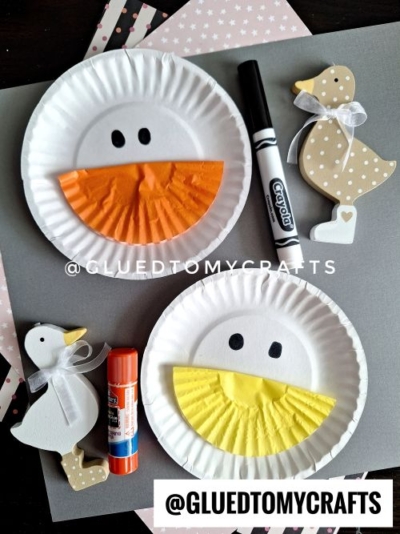A simple duck-themed craft using paper plates with black marker-drawn eyes and folded cupcake liners as beaks (one orange, one yellow). The setup includes a black marker, a red glue stick, and decorative wooden duck figures with ribbons, all arranged on a gray background with patterned paper accents. The watermark "@GLUEDTOMYCRFTS" is visible.