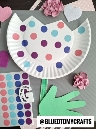 A tulip made from a white paper plate, decorated with colorful dots, has a green paper stem and hand-shaped leaf, surrounded by felt and paper hearts and flowers. @GLUEDTOMYCRAFTS is written at the bottom.