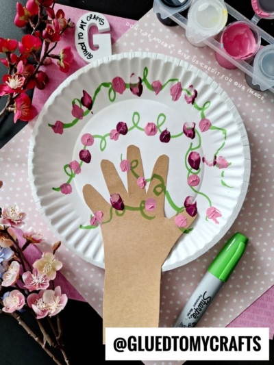 A paper plate features a hand cut-out from brown paper, glued in the center and decorated with painted pink cherry blossoms and green vines. Art supplies, flowers, and a marker surround the plate. @GLUEDTOMYCRAFTS is written at the bottom.