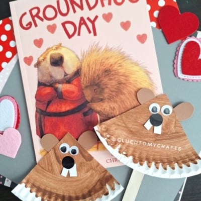 A Groundhog Day-themed craft setup featuring a children's book titled "Groundhug Day" and two paper plate groundhog crafts. The crafts are made from brown-painted paper plates with googly eyes, black noses, and white teeth, and one includes a wooden stick for holding. The scene is decorated with felt hearts and layered patterned papers in red, pink, and gray, with the watermark "@GLUEDTOMYCRAFTS" visible.
