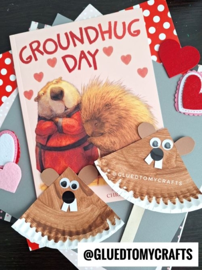 A Groundhog Day-themed craft setup featuring a children's book titled "Groundhug Day" and two paper plate groundhog crafts. The crafts are made from brown-painted paper plates with googly eyes, black noses, and white teeth, and one includes a wooden stick for holding. The scene is decorated with felt hearts and layered patterned papers in red, pink, and gray, with the watermark "@GLUEDTOMYCRAFTS" visible.