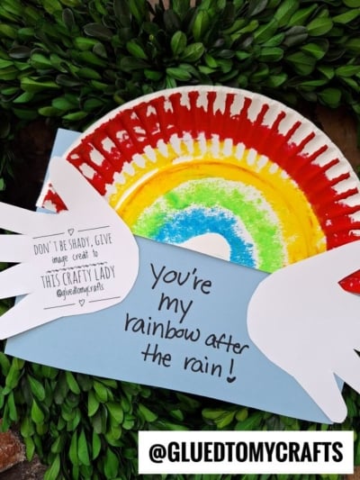A colorful paper plate rainbow craft with white paper wings and the words “You’re my rainbow after the rain!” displayed atop green foliage. A credit note and the Instagram handle @GLUEDTOMYCRAFTS are also visible.