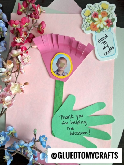 A handmade card with pink and blue paper flowers, a cut-out hand as a flower stem, and a pink flower with a child’s photo. Text reads, “Thank you for helping me blossom!” and “@GLUEDTOMYCRAFTS” at the bottom.