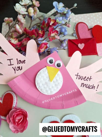 A pink paper bird craft with “I love you” and “tweet much!” written on its wings, surrounded by faux flowers, felt hearts, and a felt envelope, with the handle @GLUEDTOMYCRAFTS at the bottom.