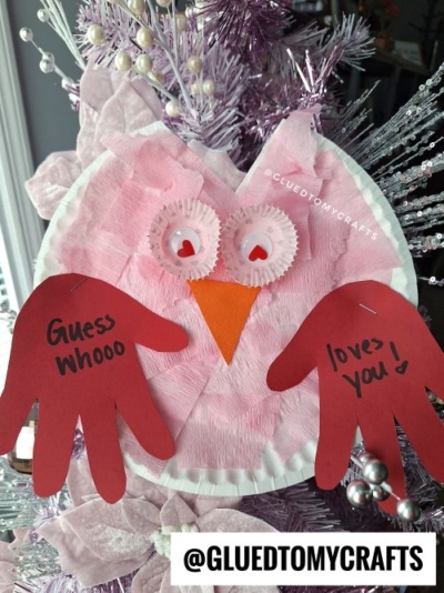 A pink owl craft made from a paper plate, crepe paper, cupcake liners, and red handprint cutouts as wings. The wings display the message "Guess Whooo loves you!" The owl's eyes feature red heart-shaped pupils, and the craft is set against a festive background of pink, purple, and silver decorative elements.