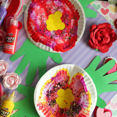 Two colorful paint splat flower crafts on white paper plates, each decorated with yellow circles in the center and glitter. The plates are placed on green paper hand cutouts, surrounded by art supplies and felt flowers.