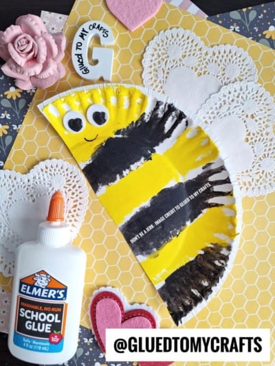 A bee craft made from a half paper plate with black and yellow stripes, googly eyes, and a smile, on a honeycomb-patterned background with heart and rose decorations, Elmers glue, and paper doilies nearby.