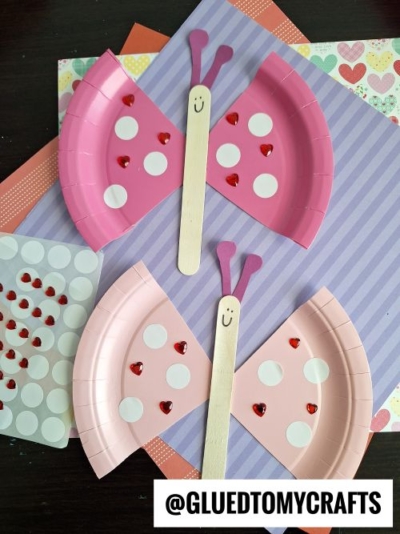 Two handmade butterfly crafts made from pink paper plates, wooden popsicle sticks, and decorative stickers (white circles and red hearts). Each butterfly has a smiley face drawn on the popsicle stick body and purple antennae. The crafts are displayed on a background of patterned papers in purple, coral, and multicolored hearts, with sticker sheets visible nearby.