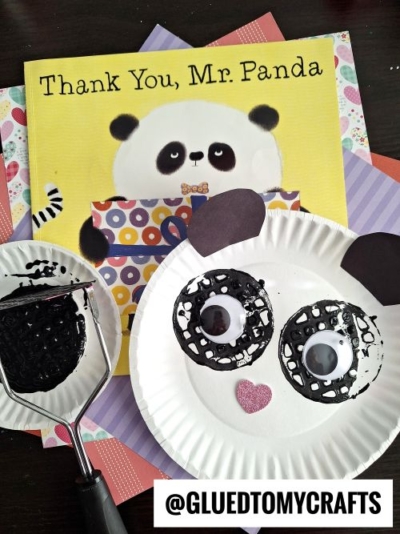 A children’s book titled “Thank You, Mr. Panda” sits behind a panda face craft made from a paper plate, black paint, and paper ears—a fun potato masher stamped craft. Art supplies and the potato masher are also visible.