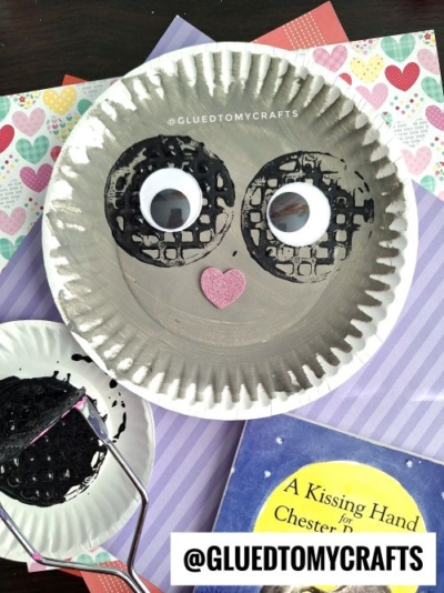 A paper plate painted gray with two large googly eyes and a pink heart-shaped nose, decorated to look like a raccoon face. Craft supplies, a children's book, and a potato masher stamped craft are nearby.