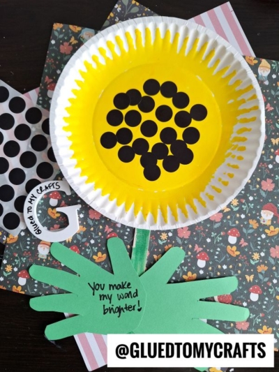 A sunflower craft made from a yellow-painted paper plate with black dot stickers in the center, green paper hand-shaped leaves, and a stem. Text on the leaf reads, You make my world brighter!.
