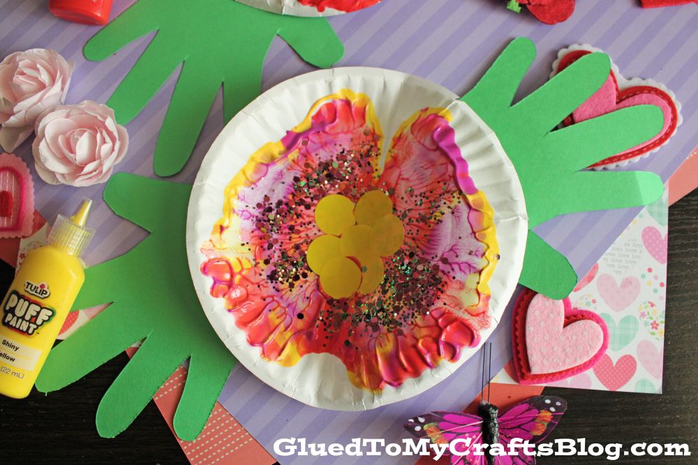 A colorful paper plate butterfly craft with yellow paint, vibrant pink and purple patterns, glitter, and green paper handprint wings, surrounded by craft supplies and decorations.