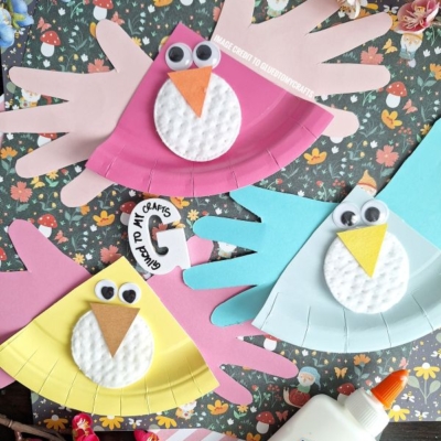 Three colorful handmade bird crafts made from paper, cotton pads, googly eyes, and orange paper beaks are displayed on a floral background with flowers, a glue bottle, and a @GLUEDTOMYCRAFTS watermark.