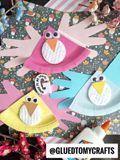 Three colorful handmade bird crafts made from paper, cotton pads, googly eyes, and orange paper beaks are displayed on a floral background with flowers, a glue bottle, and a @GLUEDTOMYCRAFTS watermark.