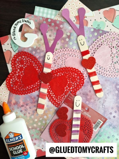 Craft supplies and finished Valentines butterfly crafts made from popsicle sticks, glitter hearts, and paper heart doily butterflies are arranged on a table with glue, scrapbook paper, and a Glued To My Crafts label visible.