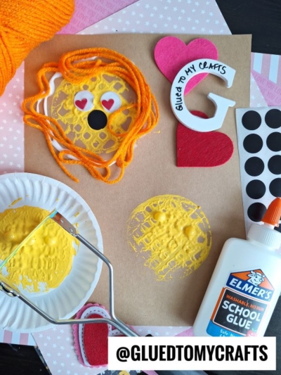 A craft scene with orange yarn, a paper plate with yellow paint, a potato masher, glue, black dot stickers, heart and letter cutouts, and a paper with a yellow paint print and a face made of yarn, googly eyes, and heart stickers.