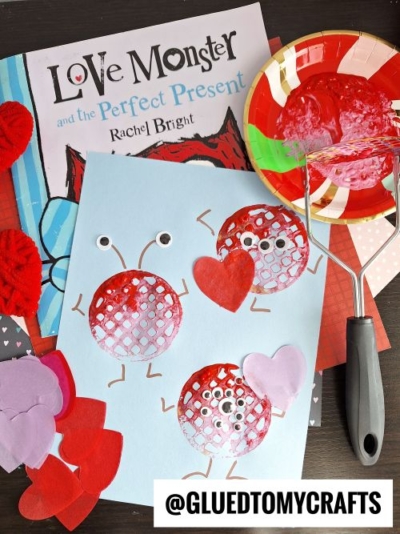 A children’s book titled “Love Monster and the Perfect Present” sits behind a valentine's day craft idea for kids—two red, textured monsters with googly eyes made using a potato masher, surrounded by colorful hearts and craft supplies.
