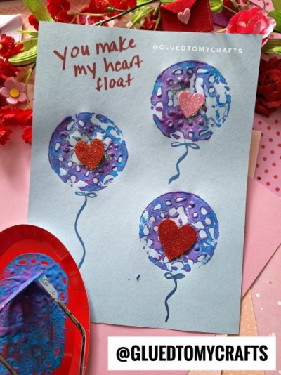 A handmade card with three purple and blue balloons, each decorated with a red glitter heart. Created using a potato masher stamped craft technique, the card says You make my heart float in red lettering and is surrounded by pink and red decor.