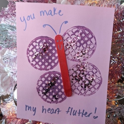 A handmade card on a pink Christmas tree shows a butterfly made from a red stick and purple doilies with glitter. The card says You make my heart flutter! and @GLUEDTOMYCRAFTS is displayed at the bottom.