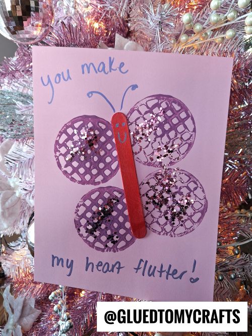 A handmade card on a pink Christmas tree shows a butterfly made from a red stick and purple doilies with glitter. The card says You make my heart flutter! and @GLUEDTOMYCRAFTS is displayed at the bottom.
