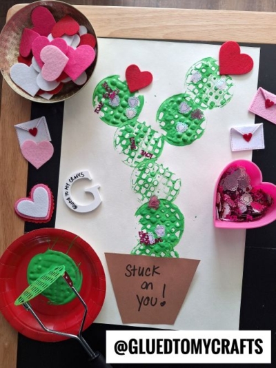 A flat lay of a Valentine's Day cactus craft on white paper. The cactus is made of green textured paint impressions, decorated with heart-shaped glitter and felt pieces. A brown paper pot at the base reads "Stuck on you!" Surrounding the craft are Valentine's-themed supplies like felt hearts, glitter, and a potato masher dipped in green paint.