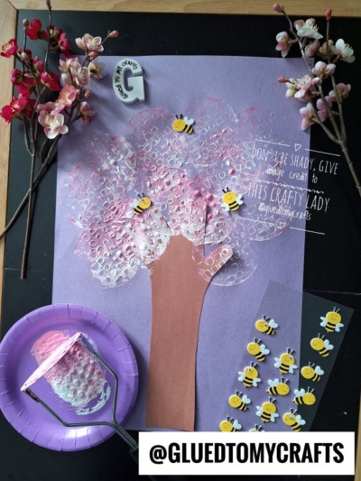 A spring craft scene features a bubble wrap-painted pink tree with a brown trunk, bee stickers, fake flowers, a purple plate, and a glue stick on a purple background. @gluedtomycrafts is written at the bottom.