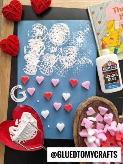 A blue paper with white paint and glue heart prints made using a potato masher stamped craft, surrounded by red yarn hearts, foam heart stickers, a glue bottle, a kids’ book, a red plate with glue, and a wooden bowl of foam hearts. @GLUEDTOMYCRAFTS is at the bottom.
