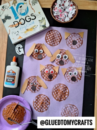 A dog-themed craft project with brown circular waffle cutouts on purple paper, decorated with googly eyes, noses, and ears. Craft supplies, a glue bottle, a bowl, and a dog book are nearby. @GLUETOMYCRAFTS.