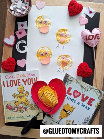 Flat lay of a Valentine's Day-themed craft setup featuring yellow duck and bird stamps made with a potato masher on white paper. The stamped designs have googly eyes, orange beaks, and some with drawn legs. Surrounding the craft are heart-shaped decorations, felt envelopes, a red heart-shaped paper plate with yellow paint and a roller, and two children's books: 'Click, Clack, Moo I LOVE YOU!' and 'LOVE finds a way.' The scene is set on a wooden and black surface with a mix of colorful craft supplies.