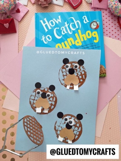 A children’s book titled “How to Catch a Groundhog” is next to a blue paper featuring three groundhog faces made from brown waffles, black and white paper, marshmallow teeth, and a fun potato masher stamped craft, surrounded by crafting supplies.