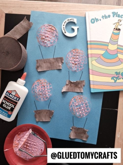 A flat lay of a hot air balloon-themed craft project on a blue sheet of paper. The craft features pink circular shapes as balloons with brown paper baskets connected by black lines. Surrounding the craft are materials like Elmer's glue, a red plate with crafting supplies, and a Dr. Seuss book titled "Oh, the Places You'll Go!" The scene is set on a dark surface with a light wooden frame.