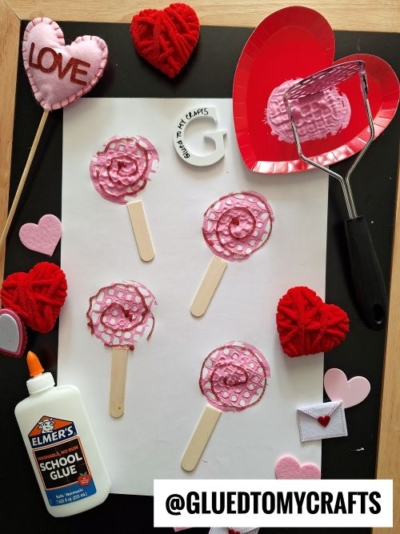"Flat lay of Valentine's Day-themed craft supplies and finished projects on a dark background with a light wooden frame. The centerpiece features pink and red 'lollipop' crafts made with textured paint and glitter on wooden sticks, arranged on white paper. Surrounding the crafts are felt hearts, a red heart-shaped paper plate with pink paint and a potato masher, a bottle of Elmer's School Glue, and a small white felt envelope with a red heart. The branding '@GLUEDTOMYCRAFTS' is visible at the bottom.