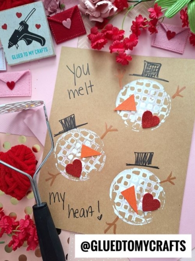 A brown card with three snowmen faces made from white paint, as part of a potato masher stamped craft, orange paper noses, and red paper hearts. The text says, You melt my heart! Surrounded by craft supplies, red flowers, and small pink envelopes.