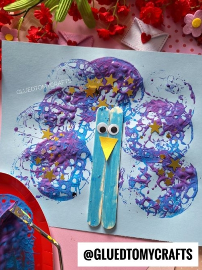 A blue and purple bubble print bird craft on light blue paper, decorated with gold star stickers, a yellow paper beak, googly eyes, and popsicle sticks for the body. Craft supplies and flowers surround this playful potato masher stamped craft.