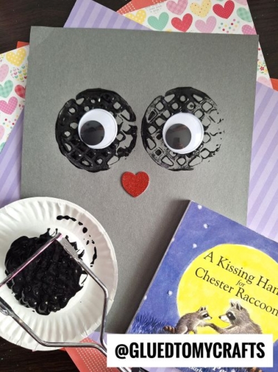 A simple raccoon-themed craft inspired by the book "A Kissing Hand for Chester Raccoon." The craft features a gray paper with black textured circles as eyes, googly eyes in the center, and a red glitter heart as the nose. A paper plate with black paint and a stamping tool are visible, along with the book and patterned papers in the background.