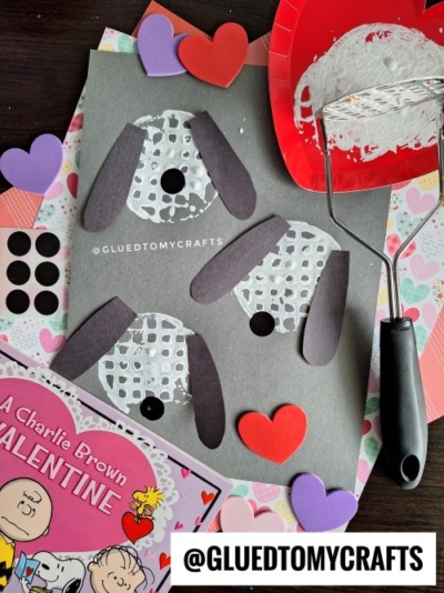 Three puppy faces made from white potato masher stamped craft circles, black paper ears, and black dot noses on gray paper. Surrounded by heart shapes, a red paper plate with white paint, and a Peanuts Valentine’s Day book.