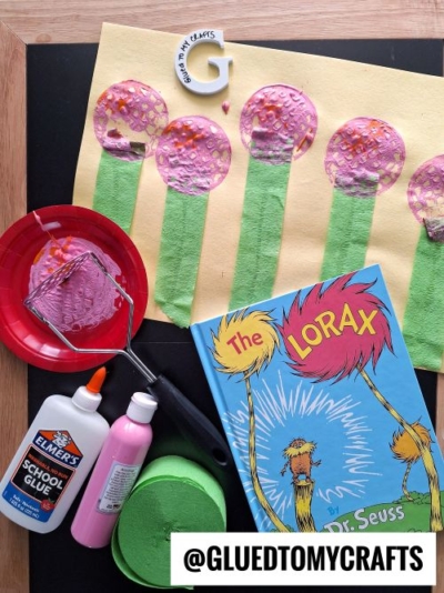 A flat lay of a craft inspired by Dr. Seuss's "The Lorax." The craft features Truffula Trees on yellow paper, with green paper trunks and textured pink and orange tops created using a potato masher. Crafting materials like pink paint, Elmer's glue, and green crepe paper are arranged around the project, along with a copy of "The Lorax" book.