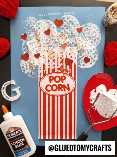 A popcorn craft for kids made with a red striped paper popcorn bag, white doilies for popcorn, red heart cutouts, gold glitter, a bottle of Elmer’s glue, and a potato masher on a blue background.